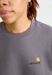 Harmaa collegepaita, jossa on ribattu kaulus ja keltainen sekä burgundinvärinen brodeerattu logo rinnassa. Pehmeä, teksturoitu kangas, rento muotoilu.