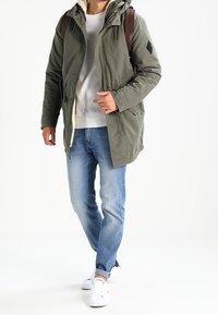 Parka con capucha de color verde oliva con cremallera, acompañada de un suéter gris claro y jeans azules de mezclilla, combinada con zapatillas blancas y una mochila marrón.