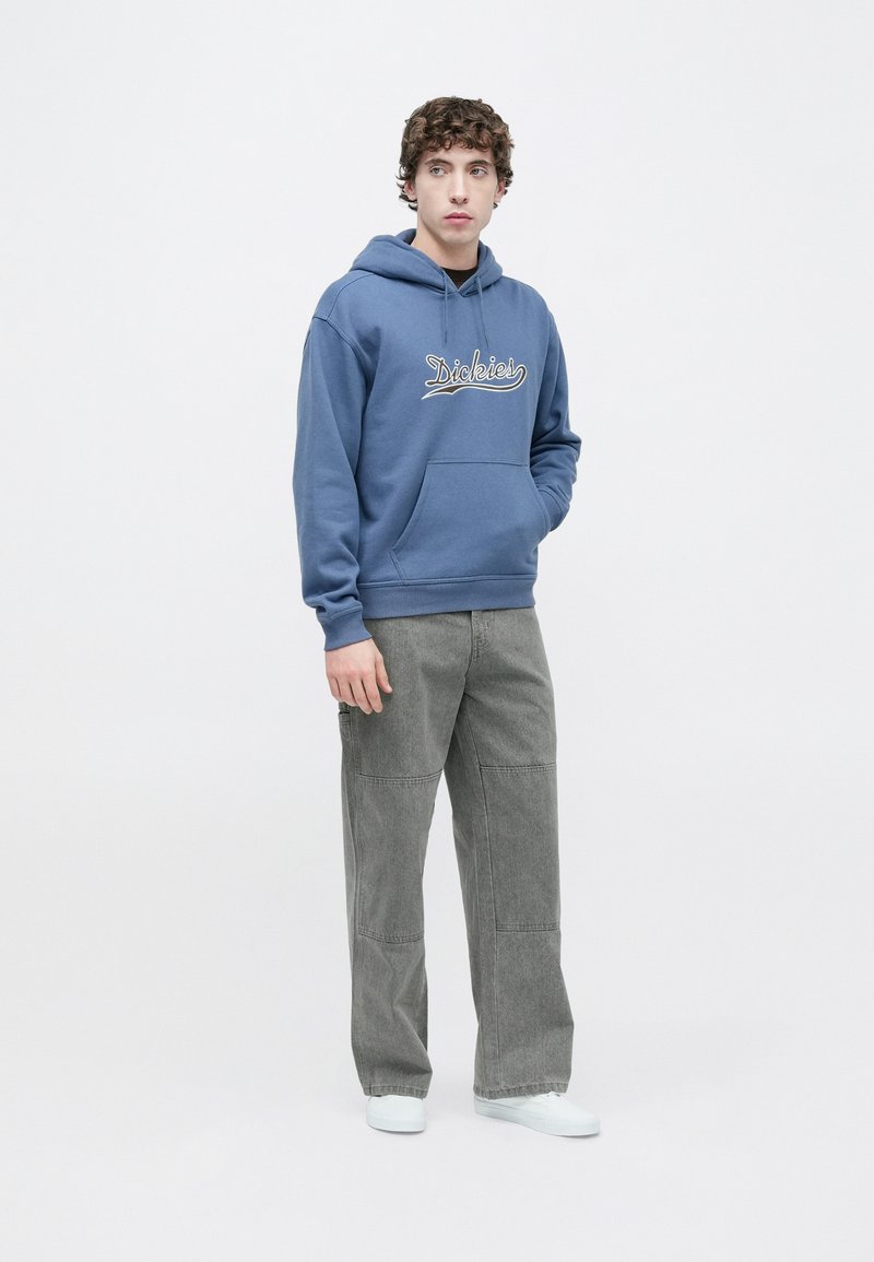 Felpa di cotone blu con tasca frontale e cappuccio con coulisse, caratterizzata da ricamo bianco "Dickies"; abbinata a pantaloni grigi a gamba larga.