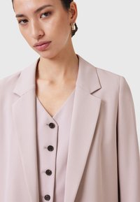 Blazer rosa chiaro con colletto a scialle, caratterizzato da bottoni neri e una texture liscia. Indossato sopra un top a scollo a V coordinato, creando un look a strati.