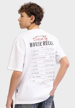 Tenåring med krøllete hår iført en hvit T-skjorte med teksten "Selskapsregler" som inkluderer spis godt, vær ærlig, lytt, dans og vær snill.