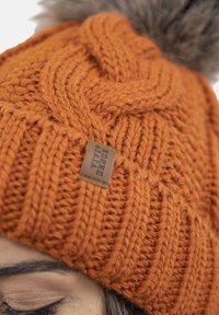 Orange strikket hue med en struktureret zigzag mønster og en falsk pels pom-pom på toppen. Inkluderer et lille læderlogo.