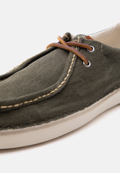 Παντόφλα από χαλαρό καμβά σε ελιά χρώμα με μπεζ ραφές, καφέ δερμάτινα κορδόνια και λευκή λαστιχένια σόλα.