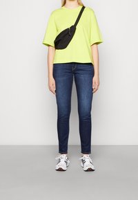Neon green oversized t-shirt, dark skinny jeans, black crossbody bag, and white sneakers with color accents. Smooth fabric, casual design.