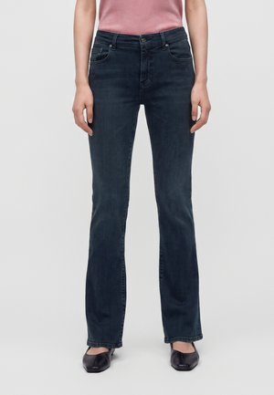 Donna che indossa jeans blu scuro a gamba dritta con tasche frontali, una maglietta rosa e scarpe nere con punta quadrata, in piedi davanti a uno sfondo semplice.