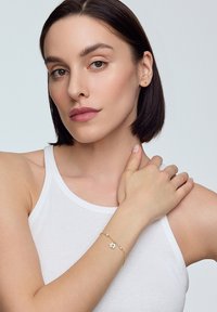 Złota bransoletka z kwiatowym wzorem, z perłowymi akcentami. Modelka ma na sobie białą koszulkę na ramiączkach, prezentując bransoletkę na swoim nadgarstku.