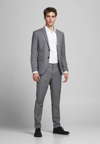 Grauer maßgeschneiderter Anzug mit Reverskragen, einreihiger Knopfschluss und schmal geschnittenen Hosen. Darunter ein weißes Hemd mit Knopfleiste und schwarze Schuhe.
