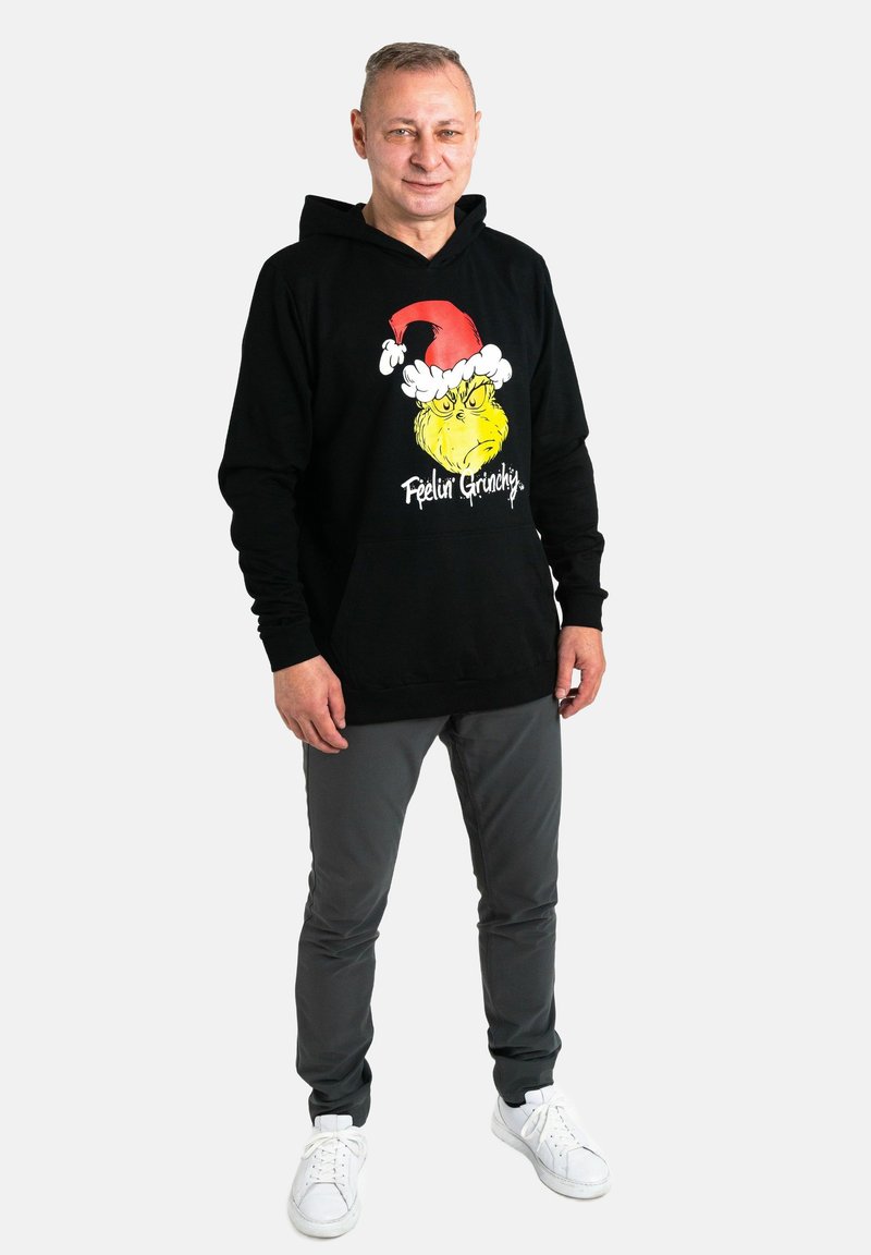 Felpa nera con un grafiche vivace del Grinch con un cappello di Babbo Natale e la scritta "Feelin' Grinchy". Abbinata a pantaloni grigi e sneakers bianche.
