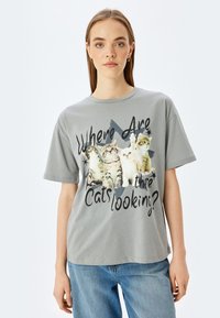 Camiseta de algodón gris que presenta un gráfico de cuatro gatos y el texto "¿A Dónde Miran los Gatos?" en negro, de corte relajado y manga corta.