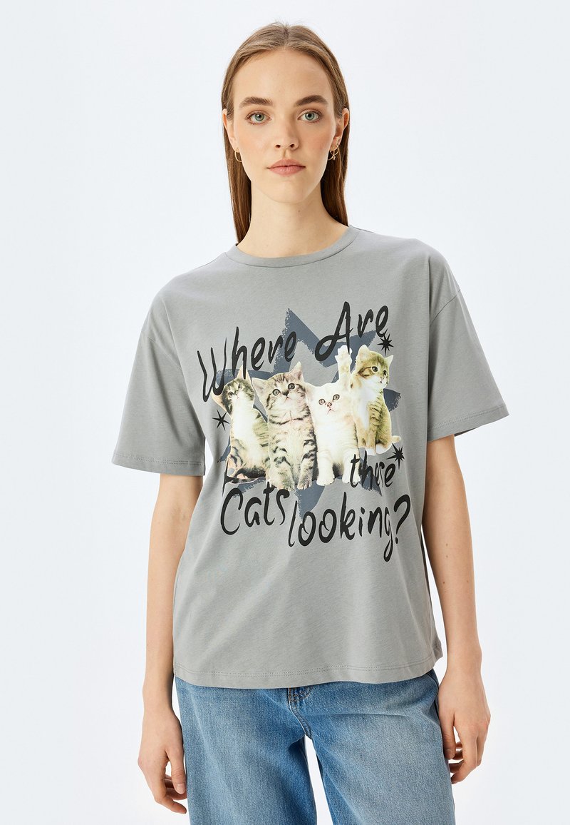 Camiseta de algodón gris que presenta un gráfico de cuatro gatos y el texto "¿A Dónde Miran los Gatos?" en negro, de corte relajado y manga corta.