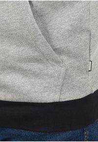 Grijze katoenen hoodie met een zak aan de linkerkant, voorzien van een zwarte geribbelde zoom. De stof heeft een gladde textuur met een lichte gemêleerde patroon.