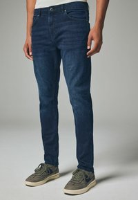 Tmavomodré denimové džínsy so slim fit strihom, s prednými vreckami, gombíkovým zapínaním a jemným vyblednutím. Skombinované so šedými teniskami.