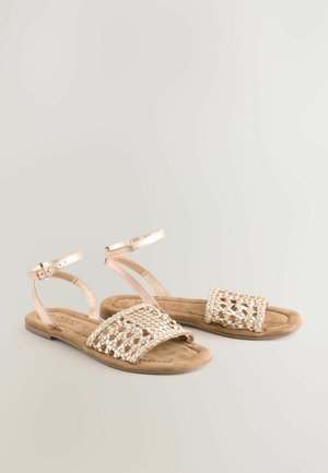 Sandalias planas con una tira metálica trenzada, suela de ante en tono claro, y correa ajustable en el tobillo, con una textura suave y diseño de punta abierta.