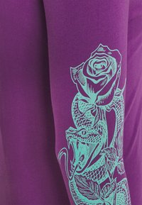 Violet pitkähihaisuus paita, jossa on turkoosivärinen kuva käärmeestä, joka on kietoutunut ruusun ympäri, esittäen monimutkaisia yksityiskohtia designissa.