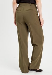 Vrouw draagt olijfgroene wijde broekspijpen en zwarte platte schoenen, staand tegen een effen witte achtergrond.