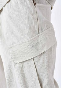 Lys beige tekstil med struktur har et syet mærke med "Project Paris" broderi. Synlige plisseringer tilføjer struktur til designet.