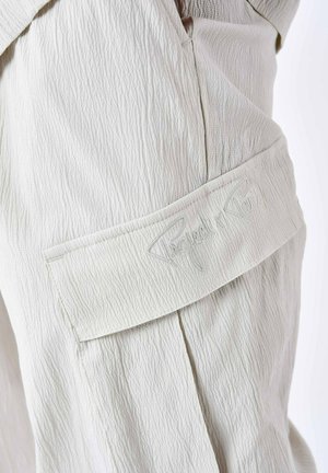 Una tela texturizada de color beige claro presenta un parche cosido con bordado de "Project Paris". Los pliegues visibles añaden estructura al diseño.