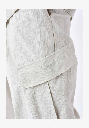 Licht beige gestructureerde stof heeft een gestikte patch met 'Project Paris' borduursel. Zichtbare plooien voegen structuur toe aan het ontwerp.