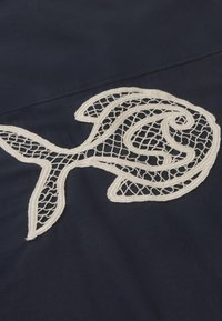 Motif de poisson blanc en dentelle brodée sur tissu foncé, présentant des motifs complexes en filet ouvert et des coutures en contour audacieuses.