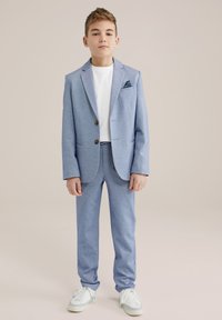 Šviesiai mėlynas džemperis ir derančios kelnės iš tekstūruoto audinio, derinamos su balta marškinėlių ir baltais sportbačiais; turi kišeninę skarelę.