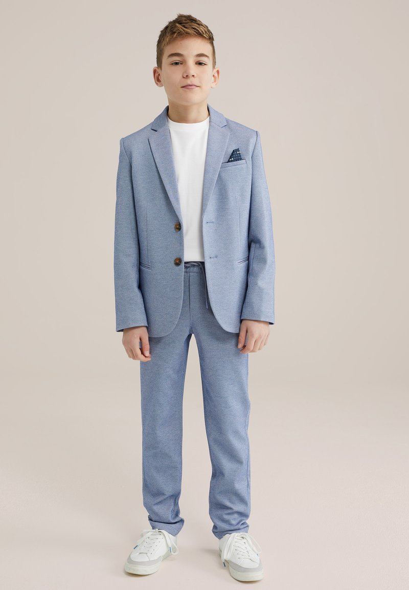 Šviesiai mėlynas džemperis ir derančios kelnės iš tekstūruoto audinio, derinamos su balta marškinėlių ir baltais sportbačiais; turi kišeninę skarelę.