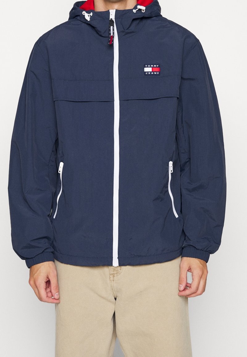 Muž v n ámořnicky modré bundě s kapucí, bílým zipem a zipy na kapsách, béžových kalhotách a malým logem Tommy Jeans na hrudi.