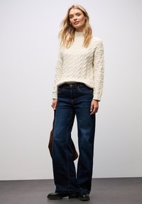 Kremowy sweter w warkocz z wysokim kołnierzem, zestawiony z ciemnoniebieskimi spodniami o szerokich nogawkach. Modelka trzyma brązową torbę, stojąc na jednolitym tle.