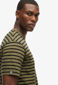 Olive green t-shirt with black horizontal stripes, featuring a round neckline and short sleeves, displaying a small logo on the sleeve.