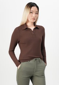 Woman with straight blonde hair wearing a brown ribbed long-sleeve polo shirt and olive green pants, hands in pockets, standing against white background.