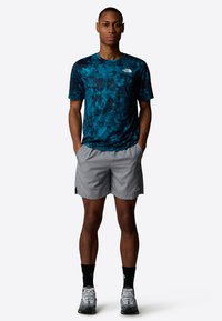 Joven de pie con las manos en los bolsillos, vistiendo una camiseta de rayas azules, pantalones cortos grises, calcetines negros y zapatillas deportivas grises, frente a un fondo liso.