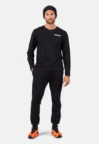 Schwarzes Langarmshirt mit kleinem weißen Logo auf der Brust, kombiniert mit schwarzen Sweatpants und leuchtend orangefarbenen Sportschuhen. Strickmütze in Schwarz.
