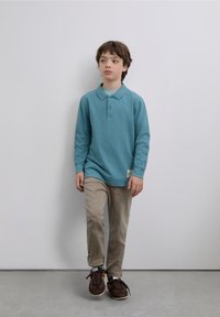 Tricou polo cu mâneci lungi de culoare turcoaz, cu guler, patru nasturi și o etichetă cusută, asortat cu pantaloni bej deschis și adidași maro.