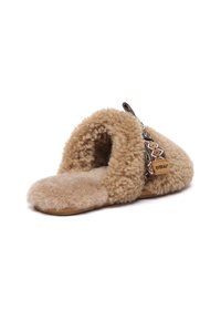 Beige slipper with soft, fuzzy texture, open back design, and a flat sole. Features a decorative strap with patterns and a brand tag.