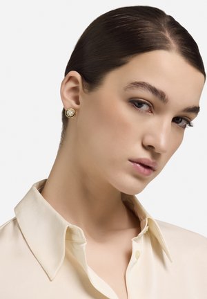 Cercei cu șurub aurii, cu un design circular texturat, având un model subtil. Purtați de un model îmbrăcat într-o bluză ușoară de mătase.