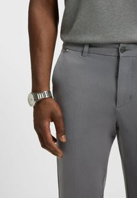 Szare dopasowane spodnie z gładkiego materiału, z prostym przodem, bocznymi kieszeniami i widocznym zapięciem na guzik. Na nadgarstku znajduje się metalowy zegarek.