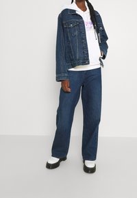 Persona vestida con una chaqueta de mezclilla oscura, sudadera blanca con impresión púrpura, pantalones anchos oscuros y botas blancas de suela gruesa frente a un fondo liso.