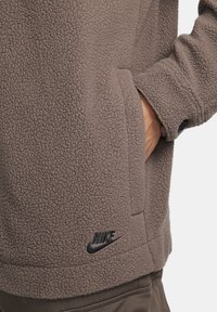 Hnědá fleecová bunda s texturovaným povrchem, vybavená bočními kapsami a černým logem Nike na spodním lemu. Detail s knoflíkem na manžetě.