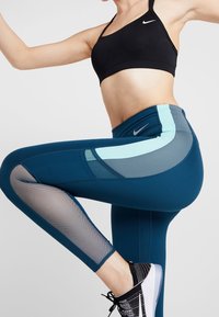 Legginsy w kolorze turkusowym z wysokim stanem, z siateczkowymi wstawkami, o zakrzywionym wzorze w kolorach jasnoniebieskim i szarym, z czarnym logo Nike na biodrze.