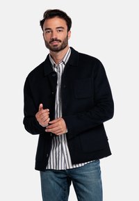 Veste en laine noire avec un design à boutons, deux poches à rabat et un col, portée sur une chemise à rayures blanches et bleues.