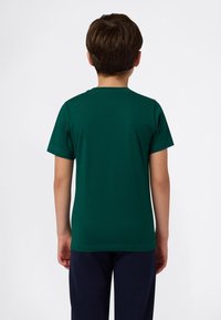 Ragazzo in piedi di spalle, con una maglietta a maniche corte verde scuro e pantaloni blu scuro, di fronte a uno sfondo bianco semplice.