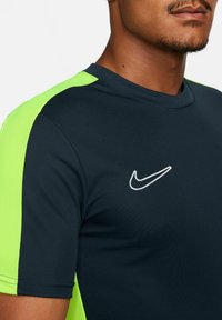 Marineblauw sportshirt met felgroene gaas zijkanten. Voorzien van een wit Nike-logo op de borst en een ronde hals. Soepele textuur.