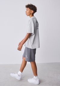 Grijs oversized t-shirt met logo, gecombineerd met grijze shorts en witte sneakers. De stof lijkt zacht, met een casual pasvorm en eenvoudig ontwerp.
