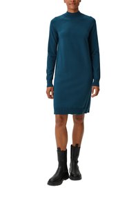 Abito lavorato a maglia color teal con collo alto, maniche lunghe e polsini a coste. Indossato con stivali neri sopra la caviglia, caratterizzati da una suola spessa e linguette per la calzata.