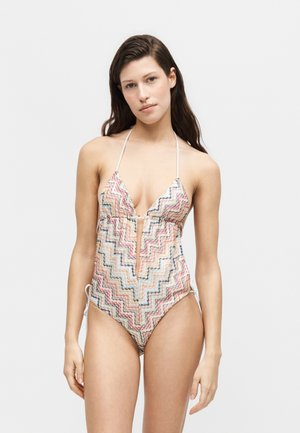 Kobieta w jednoczęściowym kostiumie kąpielowym z wielobarwnym zygzakowatym wzorem, z ramiączkami wiązanymi na szyi i wycięciem w kształcie dziurki od klucza z przodu, stojąca na jednolitym tle.