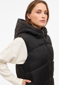 Juodas striukės tipo liemenė su gobtuvu, pasižymi siuvinėta tekstūra ir priekine užtrauktuku. Dėvima ant kreminės ilgomis rankovėmis palaidinės. Matomos šoninės kišenės.