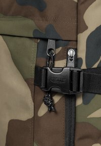 Tejido de camuflaje en tonos de verde y marrón; cuenta con una hebilla de plástico negro, cordón y cremalleras de metal. Superficie texturizada.