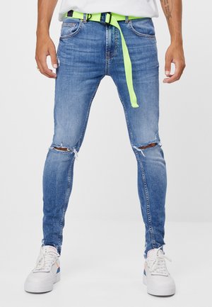Homme portant un jean skinny bleu déchiré avec des ourlets effilochés, une ceinture vert néon, des baskets blanches et une chemise blanche visible à la taille.