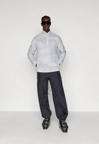 Pullover in pile blu chiaro con colletto a zip, abbinato a pantaloni impermeabili blu scuro e stivali da sci neri. Modello che indossa occhiali da sole.