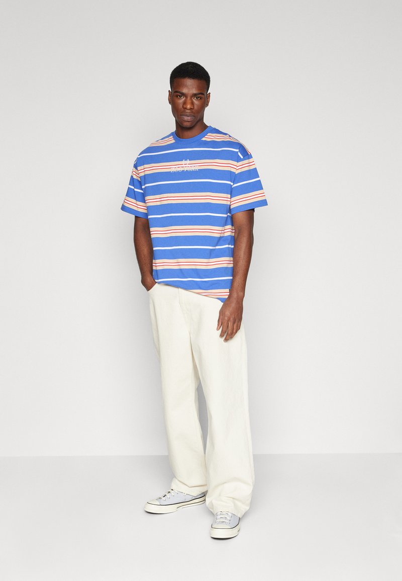 Camiseta de rayas azules con rayas naranjas y blancas, cuello redondo, mangas cortas, corte casual, combinada con pantalones de pierna ancha en color crema y zapatillas ligeras.