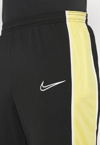 Schwarze Sportshorts mit gelben Seitenstreifen. Sie verfügen über einen elastischen Bund und ein weißes gesticktes Logo auf der linken Seite. Texturiertes Material.